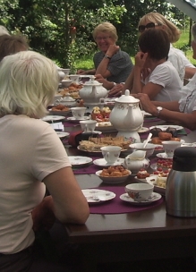 Groep mensen geniet buiten in de boomgaard aan tafel van theeleut hightea met diverse huisgemaakte zoete en hartige lekkernijen